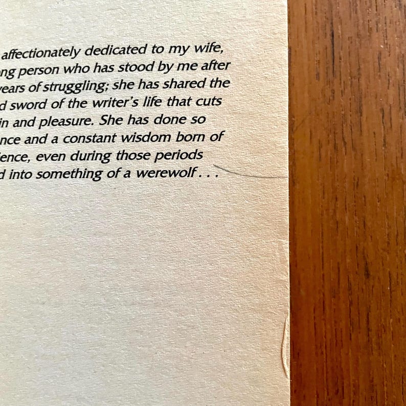 May include: Close-up of a book page with printed text. The text is in black font on a cream-colored paper. The right side of the image shows a wooden surface. The text includes phrases like "dedicated to my wife" and "something of a werewolf."