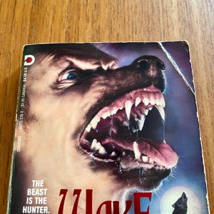 May include: A book cover with a close-up of a snarling dog's face with sharp teeth. The cover has a dark blue and purple background with the text "THE BEAST IS THE HUNTER." and the title "Zlave".