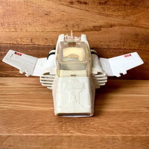 May include: A white toy spaceship with wings and a clear cockpit. The toy has red accents on the wings and is set against a wooden background. The wings are extended, and the toy appears to be a vintage collectible.