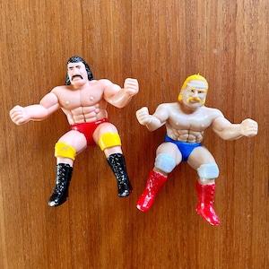 May include: Two vintage wrestling action figures. One figure wears red trunks, yellow knee pads, and black boots. The other figure wears blue trunks, white knee pads, and red boots. Both figures have muscular physiques and are posed in a fighting stance.