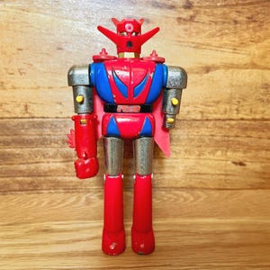 May include: A vintage robot toy with a red, blue, and silver color scheme. The robot has a red head with horns, a blue chest with red accents, and silver arms and legs. It has a black belt and red cape.