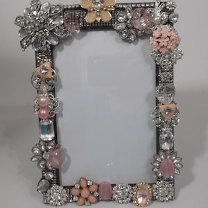 May include: A silver picture frame decorated with pink and clear rhinestones, faux pearls, and various vintage brooches. The frame is adorned with a variety of floral and geometric designs.