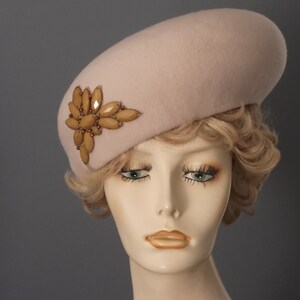 May include: A light pink felt beret with a large, ornate, gold-toned embellishment on the side.