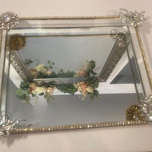 May include: Rectangular mirrored tray with a gold beaded border and ornate silver corner embellishments. The tray features floral accents and decorative brooches. A vintage-inspired decorative piece.