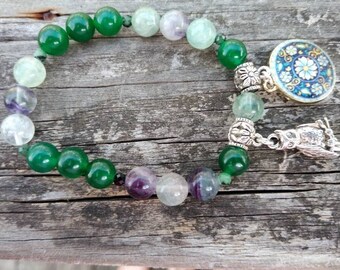May include: A green and purple gemstone bracelet with a silver owl charm and a round blue and white patterned charm.