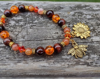 May include: A beaded bracelet featuring a mix of orange, brown, and gold beads. The bracelet has a gold sunflower charm and a gold bee charm. The beads are various sizes and shapes, creating a unique accessory.