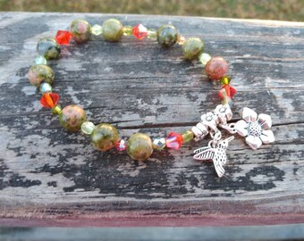 May include: A green, red, and clear gemstone bracelet with a silver hummingbird and flower charm.