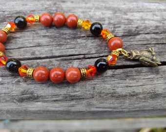 May include: A beaded bracelet featuring reddish-orange and black beads, accented with gold-colored spacers and orange and yellow crystals. A small, bronze-colored owl charm hangs from the bracelet. The bracelet is displayed on a weathered wooden surface.