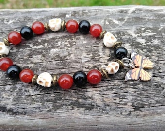 May include: A beaded bracelet featuring red, black, and off-white skull beads, interspersed with red beads. A butterfly charm with orange and black wings dangles from the bracelet. The bracelet is displayed on a weathered wooden surface.