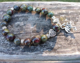 May include: A beaded bracelet featuring green and burgundy stone beads. The bracelet has silver-toned spacers and a dragon charm. The beads are round and vary in size. The bracelet is displayed on a weathered wooden surface.