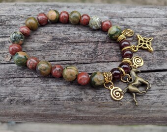 May include: A beaded bracelet featuring a mix of red, green, and tan stone beads. Gold-colored charms include a horse, a Celtic knot, and spiral designs. The bracelet is displayed on a wooden surface.