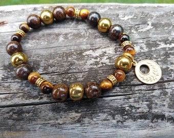 May include: A brown and gold beaded bracelet with a gold charm featuring a wreath design. The bracelet is made with tiger's eye, hematite, and wood beads.