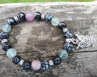 May include: A black, white, and blue beaded bracelet with an owl charm. The beads are made of snowflake obsidian, amethyst, and fluorite.