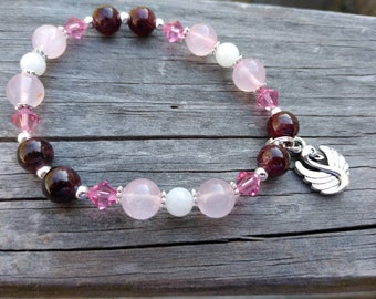 May include: A pink, white, and burgundy beaded bracelet with a silver swan charm. The bracelet is made with rose quartz, moonstone, and garnet beads.