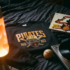 Könnte beinhalten: Schwarzes Sweatshirt mit "PIRATES" und einer Baseballspieler-Grafik. Ein Baseball und eine Vintage-Schallplatte liegen auf einer dunkelgrauen Bettdecke. Eine Lavalampe befindet sich im Hintergrund.