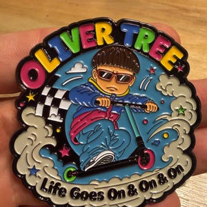 May include: A circular enamel pin featuring a cartoon character with a bowl cut and sunglasses riding a scooter. The text OLIVER TREE is at the top, and Life Goes On & On & On is at the bottom. The design includes a checkered flag and clouds.
