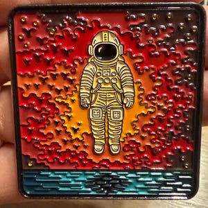 May include: A square enamel pin featuring a central cream-colored astronaut in a space suit against a vibrant background of red, orange, and yellow cloud-like shapes. Below the astronaut is a horizontal band of textured, dark teal horizontal lines.