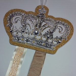 May include: A gold crown with white and silver details, adorned with faux pearls and rhinestones. Two ribbons, one white with gold accents and one brown, hang from the bottom of the crown.