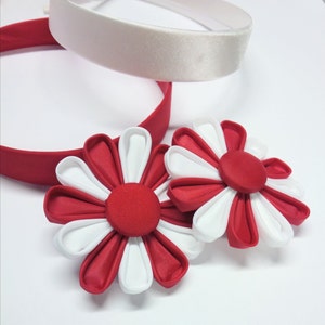 May include: Two red and white fabric flowers with a center button. The flowers are attached to a red headband with a white satin headband underneath.