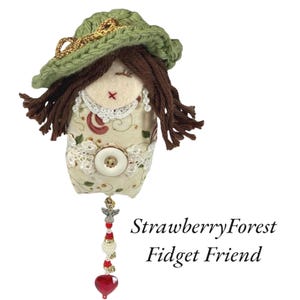 May include: A handmade fabric doll with brown yarn hair, a green crocheted hat, and a decorative beaded charm. The doll's body is made of patterned fabric with lace and a large button. The text "StrawberryForest Fidget Friend" is visible.
