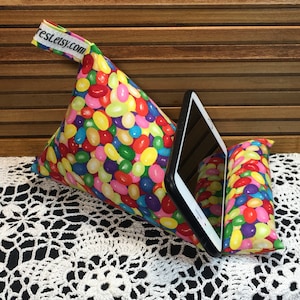 May include: A colorful bean-shaped pillow with a tablet stand. The pillow is made of fabric with a pattern of multi-colored jelly beans. The tablet stand is made of a solid color fabric. The pillow is on a white crocheted surface. The text "rest.etsy.com" is on the pillow.