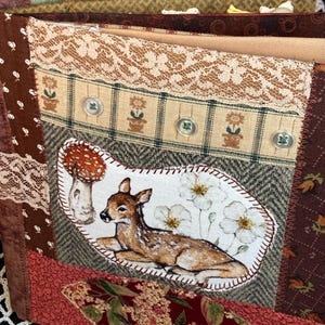 May include: A handmade fabric book with a patchwork design. The cover features a deer, mushrooms, and flowers. The book is decorated with lace, buttons, and various patterned fabrics.