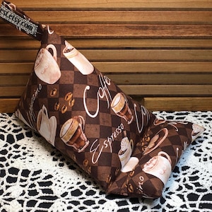 Puede incluir: Una almohada de cuña marrón y blanca con estampado y asa. La almohada tiene un tema de café con imágenes de tazas de café, granos de café y las palabras "Coffee", "Espresso" y "Cappuccino".