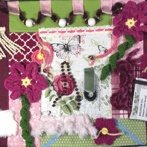 May include: A sensory activity mat featuring a burgundy ribbed border, a central panel with butterfly and rose prints, and various tactile elements including crocheted magenta flowers, white fringe, faux fur, beads, and a pocket labeled Belongs to.