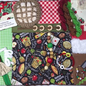 May include: A sensory activity mat with various textures and elements. Features a crocheted item, wooden rings, fabric with apple and pie patterns, a zipper, and a small tag that reads "LONGS TO: Insert your loved one's same in pocket."