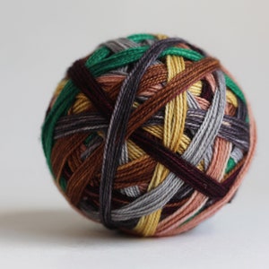 Op de afbeelding: Een bolvormige kluwen garen gewikkeld met meerdere draden in diverse kleuren, waaronder donkergroen, mosterdgeel, lichtgrijs, donkerbruin en gedempt roze, wat zorgt voor een getextureerd, verweven oppervlak met een dicht, gelaagd uiterlijk.