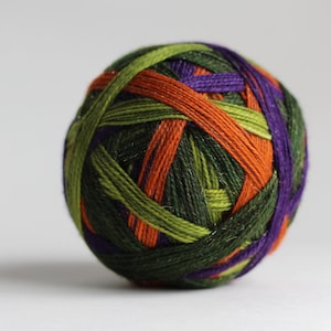 Puede incluir: Una bola esférica de hilo enrollada con múltiples hebras en tonos verde bosque oscuro, verde lima brillante, naranja vibrante y morado intenso. Las fibras están apretadamente enrolladas para crear un objeto redondo, texturizado y multicolor.