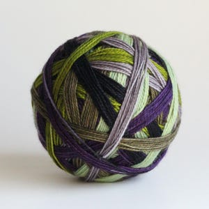 May include: A spherical ball of yarn wound with multiple strands of thread in varying shades of deep purple, olive green, lime green, black, and light gray. The fibers are tightly coiled, creating a textured, multi-colored surface pattern.