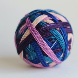 Op de afbeelding: Een veelkleurige bol wol, met tinten blauw, paars, roze en perzik, die door elkaar zijn geweven. Het garen is strak opgerold tot een bolvorm. Geschikt voor brei- of haakprojecten.