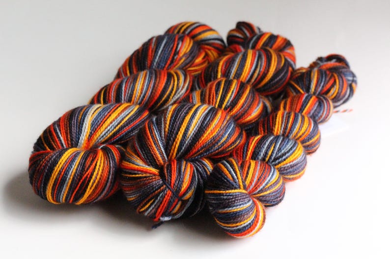 May include: A collection of colorful yarn skeins. The yarn features a striped pattern with shades of orange, yellow, navy blue, and gray. The skeins are arranged in a cluster, showcasing the texture and color variations of the yarn. Ideal for knitting and crochet projects.
