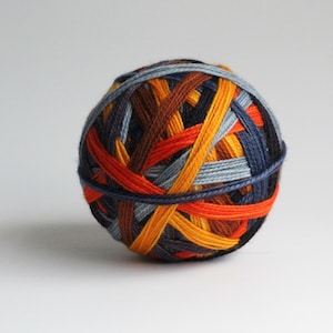May include: A ball of yarn with a colorful, woven design. The yarn features strands of orange, yellow, brown, blue, and gray, creating a textured, spherical shape. The yarn is suitable for knitting or crocheting projects.