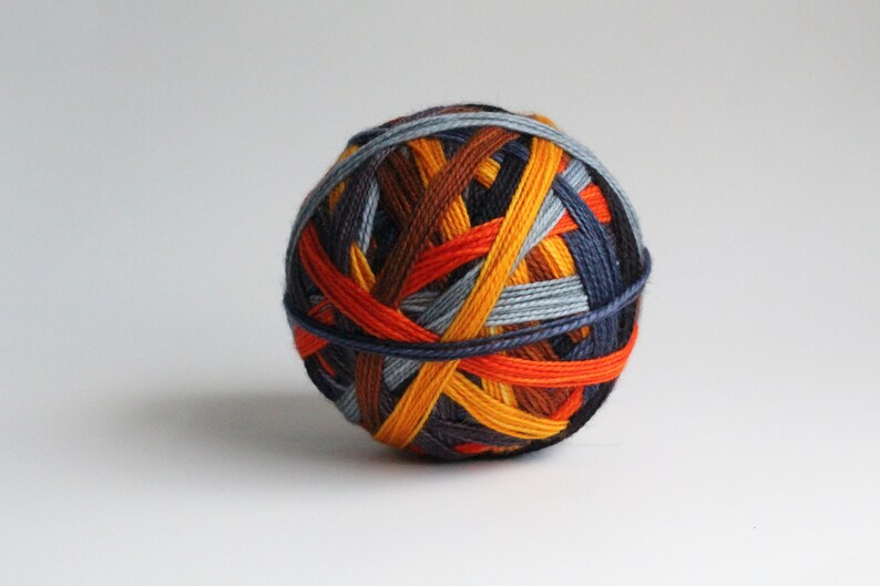 May include: A ball of yarn with a colorful, woven design. The yarn features strands of orange, yellow, brown, blue, and gray, creating a textured, spherical shape. The yarn is suitable for knitting or crocheting projects.