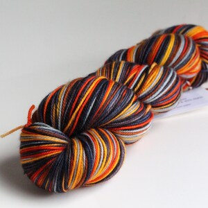 May include: A skein of colorful yarn with a mix of orange, yellow, navy blue, and gray stripes. The yarn is tightly wound and has a tag attached. This yarn is suitable for knitting or crocheting projects.