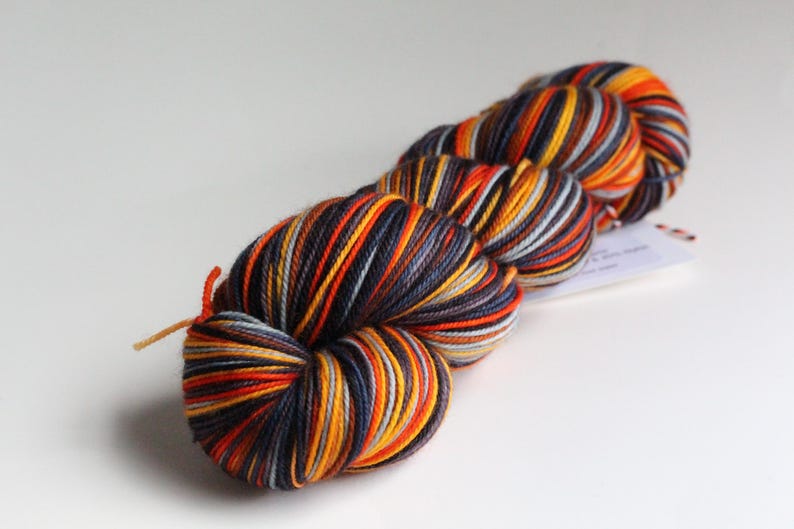 May include: A skein of colorful yarn with a mix of orange, yellow, navy blue, and gray stripes. The yarn is tightly wound and has a tag attached. This yarn is suitable for knitting or crocheting projects.