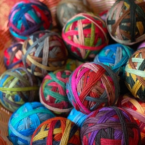 May include: A collection of colorful yarn balls in a woven basket. The yarn balls are a mix of vibrant colors, including blues, greens, reds, and purples, creating a visually appealing display. The yarn is tightly wound, and the basket is a light pink color.