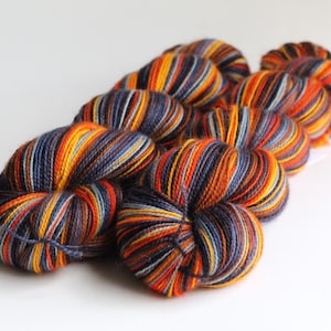 May include: A collection of colorful yarn skeins. The yarn is a mix of orange, yellow, blue, and purple, creating a striped pattern. The skeins are arranged in a diagonal line against a white background, ideal for knitting or crochet projects.
