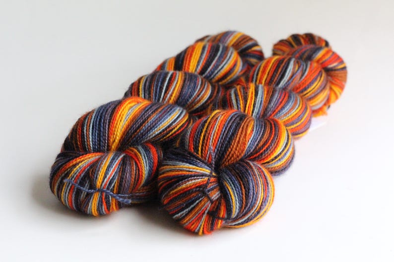 May include: A collection of colorful yarn skeins. The yarn is a mix of orange, yellow, blue, and purple, creating a striped pattern. The skeins are arranged in a diagonal line against a white background, ideal for knitting or crochet projects.