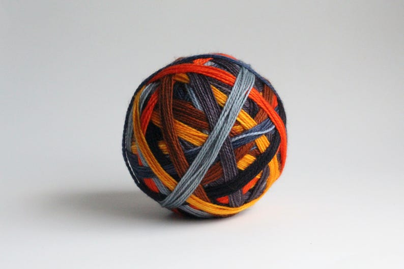 May include: A ball of colorful yarn with a mix of orange, yellow, brown, blue, and gray strands. The yarn is tightly wound, creating a textured, spherical shape. Ideal for knitting or crochet projects.