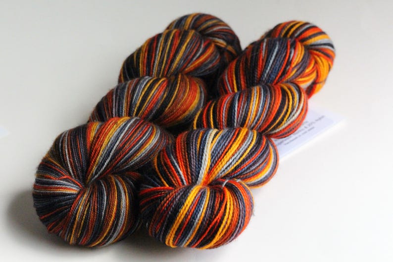 May include: A collection of colorful yarn skeins. The yarn features a striped pattern with shades of orange, yellow, black, and gray. The yarn is arranged on a white surface, showcasing the vibrant colors and textures.