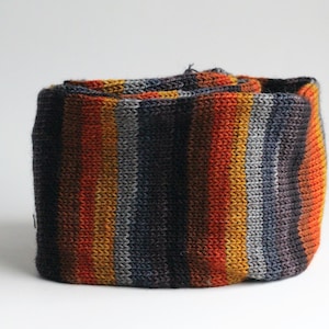 May include: A rolled-up, knitted scarf with vertical stripes in shades of orange, yellow, gray, and navy blue. The scarf appears to be made of a soft, textured yarn. The color palette is warm and autumnal.