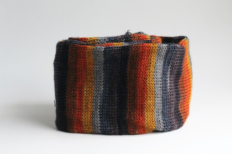 May include: A rolled-up, knitted scarf with vertical stripes in shades of orange, yellow, gray, and navy blue. The scarf appears to be made of a soft, textured yarn. The color palette is warm and autumnal.