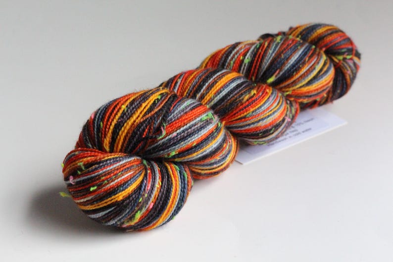 May include: A skein of colorful yarn with orange, yellow, gray, and navy blue strands. The yarn has a speckled appearance with small flecks of green. The yarn is likely used for knitting or crocheting projects.