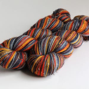 May include: Close-up of several skeins of yarn. The yarn is a mix of colors, including orange, yellow, red, gray, and white. The yarn is arranged in a loose, coiled shape, and the colors are striped throughout the skeins. The background is plain white.