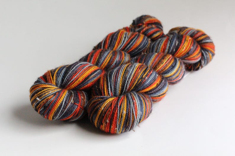 May include: Close-up of several skeins of yarn. The yarn is a mix of colors, including orange, yellow, red, gray, and white. The yarn is arranged in a loose, coiled shape, and the colors are striped throughout the skeins. The background is plain white.