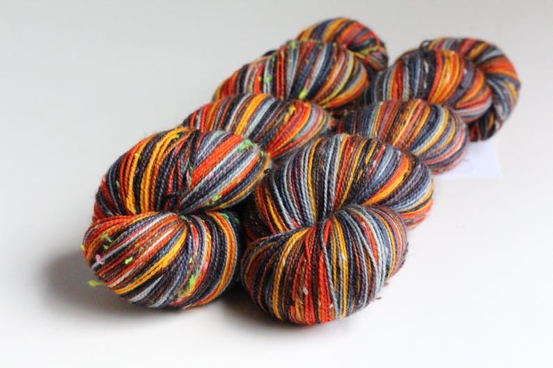 May include: A collection of colorful yarn skeins. The yarn features a mix of orange, yellow, gray, and black strands, creating a textured appearance. The skeins are arranged in a cluster, showcasing the vibrant color palette and potential for crafting projects.