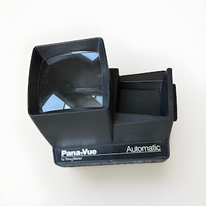 May include: A black Pana-Vue by View-Master Automatic slide viewer featuring a large rectangular magnifying lens on the left side and a slot for inserting slides on the right. The device has a matte finish with white text printed on the front.